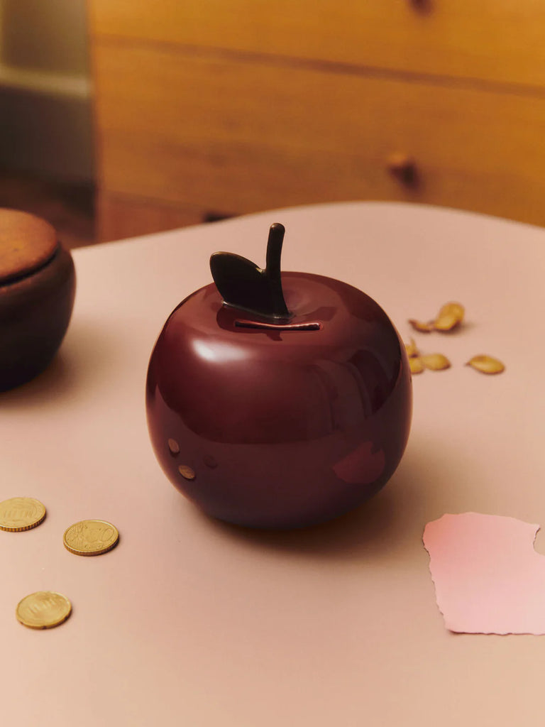 A Ferm Living Apple Money Bank, deep red and glossy with a top coin slot, rests on a light table amid scattered coins and a torn pink paper. Ideal for kids' rooms, it pops against a softly blurred wooden dresser. Ships in one week.