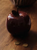A glossy, dark red Ferm Living Apple Money Bank with a stem top rests on a wooden surface beside gold coins—perfect as a ceramic piggy bank or charming kids’ room decor. Ships in one week.