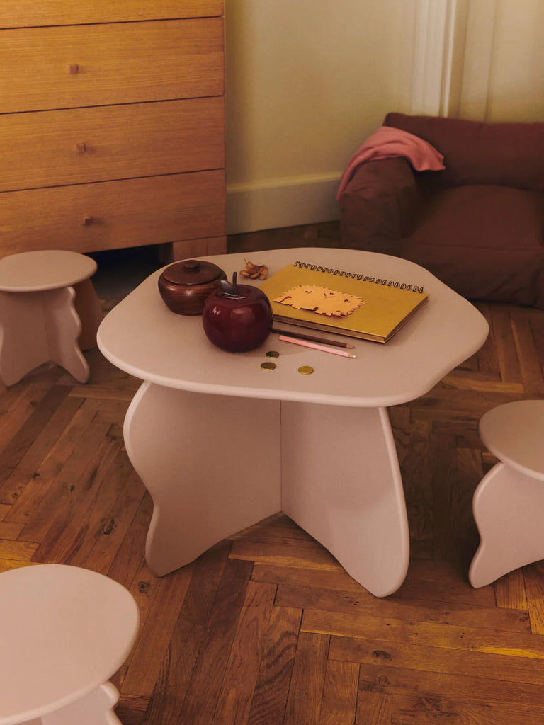 A small beige kids’ table with art supplies, apple, bowl, and the Ferm Living Apple Money Bank (ships in one week) on top, surrounded by matching stools—perfect for children’s room decor with a dresser and cushioned chair nearby.