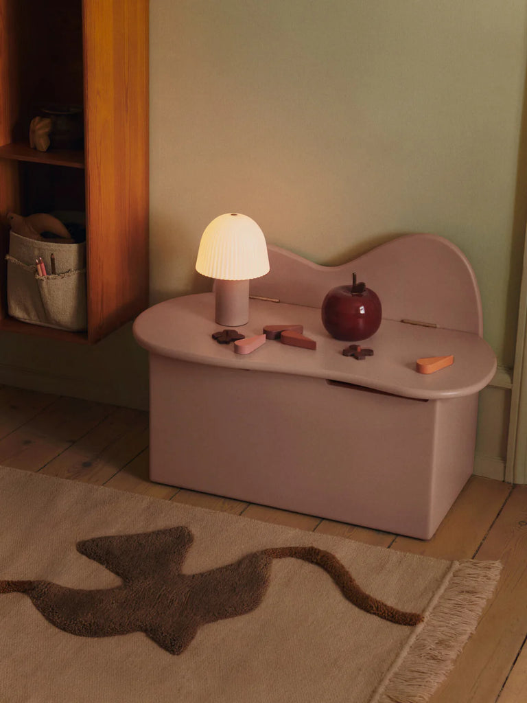 A small mauve storage bench against a green wall holds a ribbed white lamp, the Ferm Living Apple Money Bank (ships in one week), and several wooden toys. A beige rug with a brown abstract pattern covers the wooden floor.