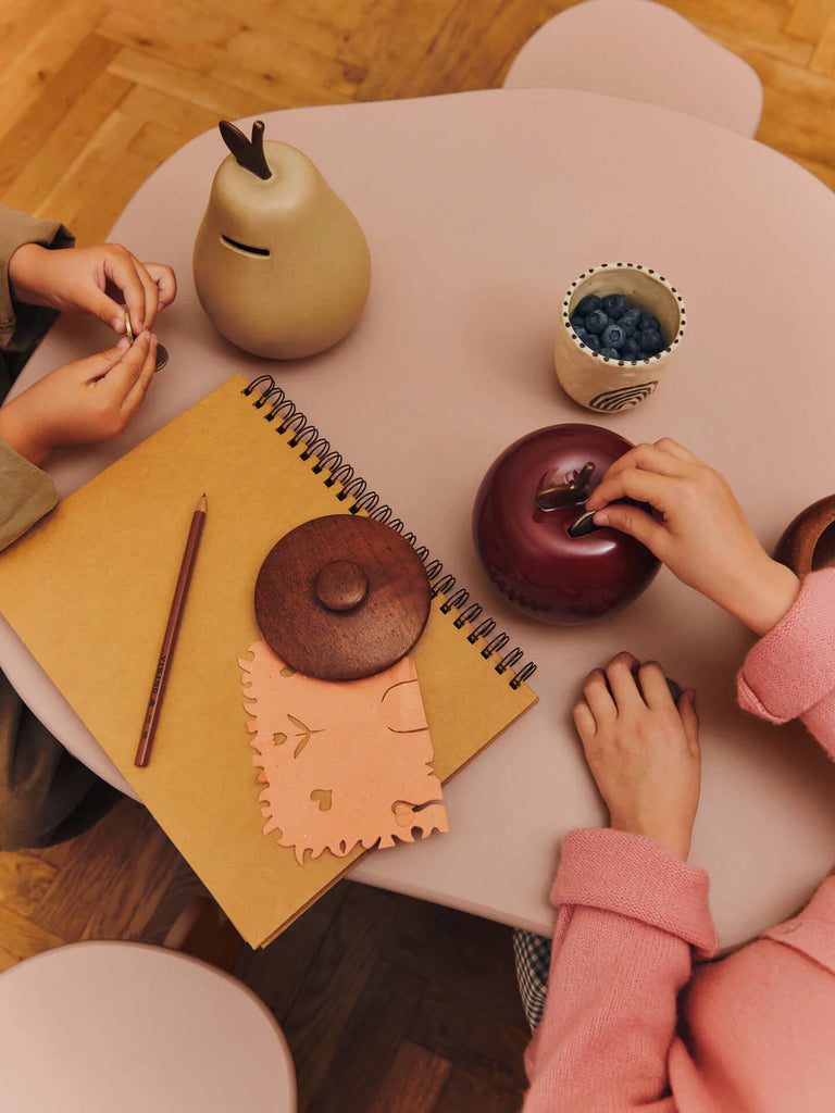 Two children’s hands rest on a small pink table with a spiral notebook, pencil, cup of blueberries, cloth, and the Ferm Living Apple Money Bank—ships in one week—perfect as kids room decor, all neatly arranged on the tabletop.