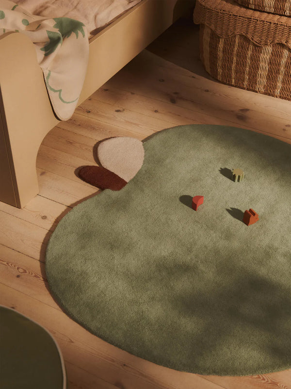 The Ferm Living Apple Tufted Rug - Tea Green, shaped like an apple with brown and cream accents, rests on a light wooden floor by a woven basket and bed. Made from hand-tufted New Zealand wool, it's ideal for playrooms with colorful blocks scattered on top.