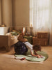 A young child balances on a star-patterned exercise ball, drawing on a notepad atop the Ferm Living Apple Tufted Rug - Tea Green in a cozy bedroom filled with sunlight, a small bed, stuffed toy, and woven baskets.