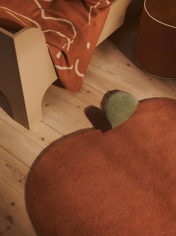 A cozy room with light wooden flooring, a beige bed with an orange patterned blanket, and the Ferm Living Apple Tufted Rug - Brown Clay with a green leaf beside a round brown side table—perfect as a playful kids' playroom rug.