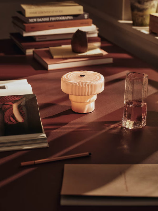 A sunlit desk with stacked books, a glass of water, a closed envelope, a pen, and the Ferm Living Ripple Portable Lamp - Pale Rose—a round, ribbed dimmable LED lamp—at the center amid warm light and soft shadows.