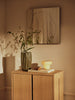 A minimalist wooden cabinet with a green vase of leafy branches, stacked books, the Ferm Living Ripple Portable Lamp - Oat with dimmable LED light, and abstract wall art above—all bathed in warm sunlight.