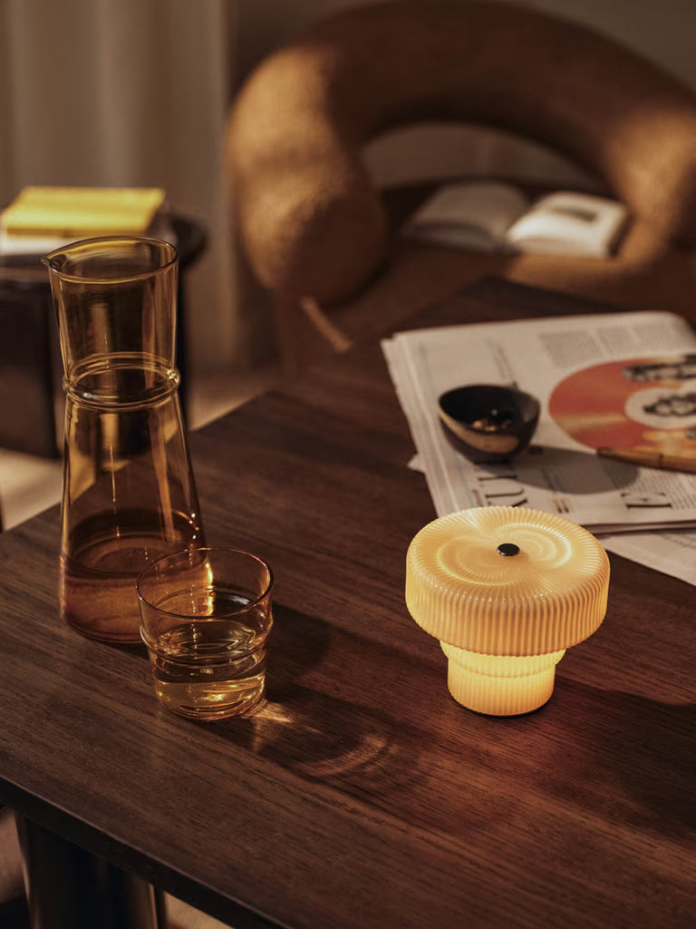 The Ferm Living Ripple Portable Lamp – Oat sits with an amber glass carafe, matching tumbler, and bowl on a dark wood table scattered with newspapers and a book, next to a curved brown armchair in a cozy setting.