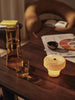 The Ferm Living Ripple Portable Lamp – Oat sits with an amber glass carafe, matching tumbler, and bowl on a dark wood table scattered with newspapers and a book, next to a curved brown armchair in a cozy setting.