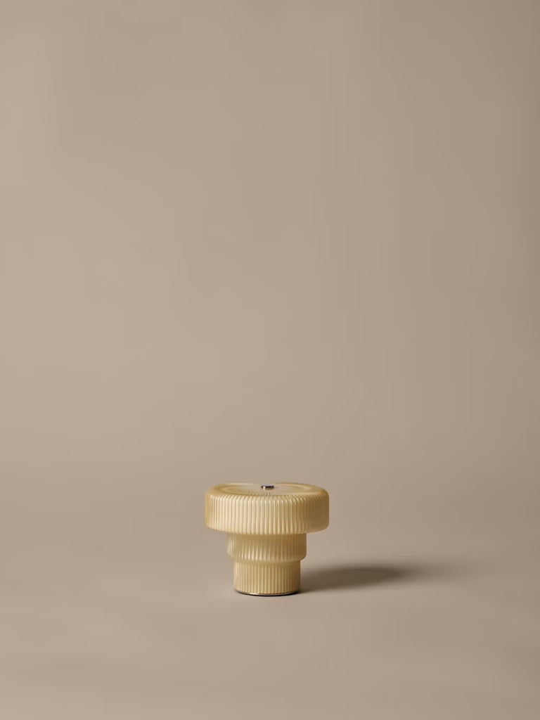 A small, beige, ribbed plastic screw cap stands upright on a plain light brown surface with a minimal background—echoing the understated elegance of the Ferm Living Ripple Portable Lamp in Oat.