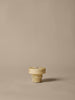 A small, beige, ribbed plastic screw cap stands upright on a plain light brown surface with a minimal background—echoing the understated elegance of the Ferm Living Ripple Portable Lamp in Oat.