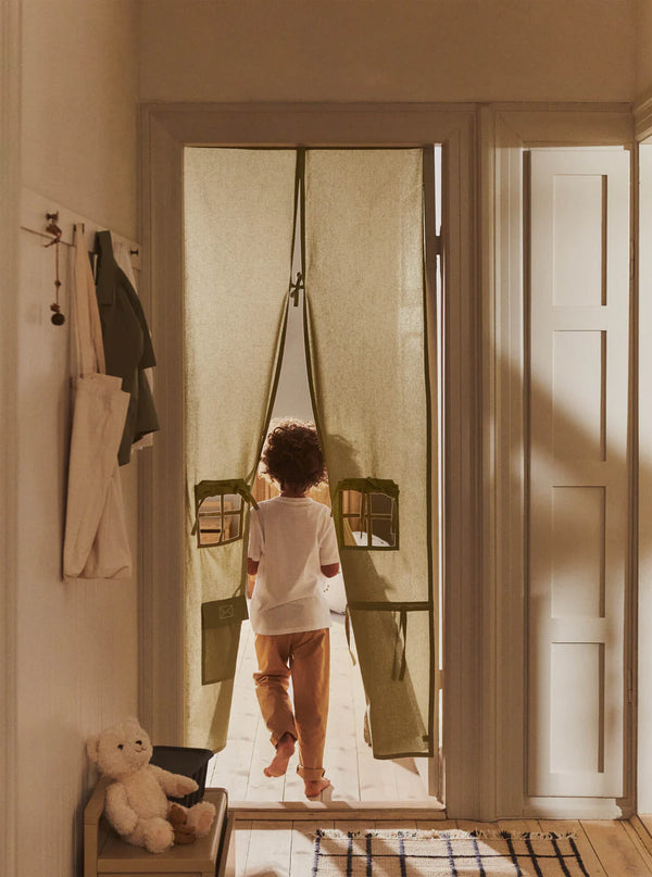 A child walks barefoot through a doorway adorned with the Ferm Living Settle Door Curtain in Elm Green, crafted from recycled cotton and featuring two small window cutouts. A white teddy bear sits on a bench nearby, as sunlight streams into the cozy hallway.
