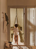 A child walks barefoot through a doorway adorned with the Ferm Living Settle Door Curtain in Elm Green, crafted from recycled cotton and featuring two small window cutouts. A white teddy bear sits on a bench nearby, as sunlight streams into the cozy hallway.