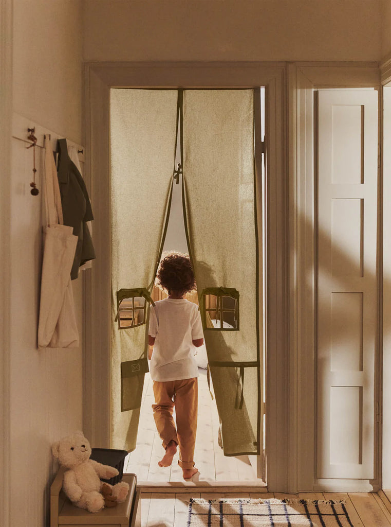 A child walks barefoot through a doorway adorned with the Ferm Living Settle Door Curtain in Elm Green, crafted from recycled cotton and featuring two small window cutouts. A white teddy bear sits on a bench nearby, as sunlight streams into the cozy hallway.