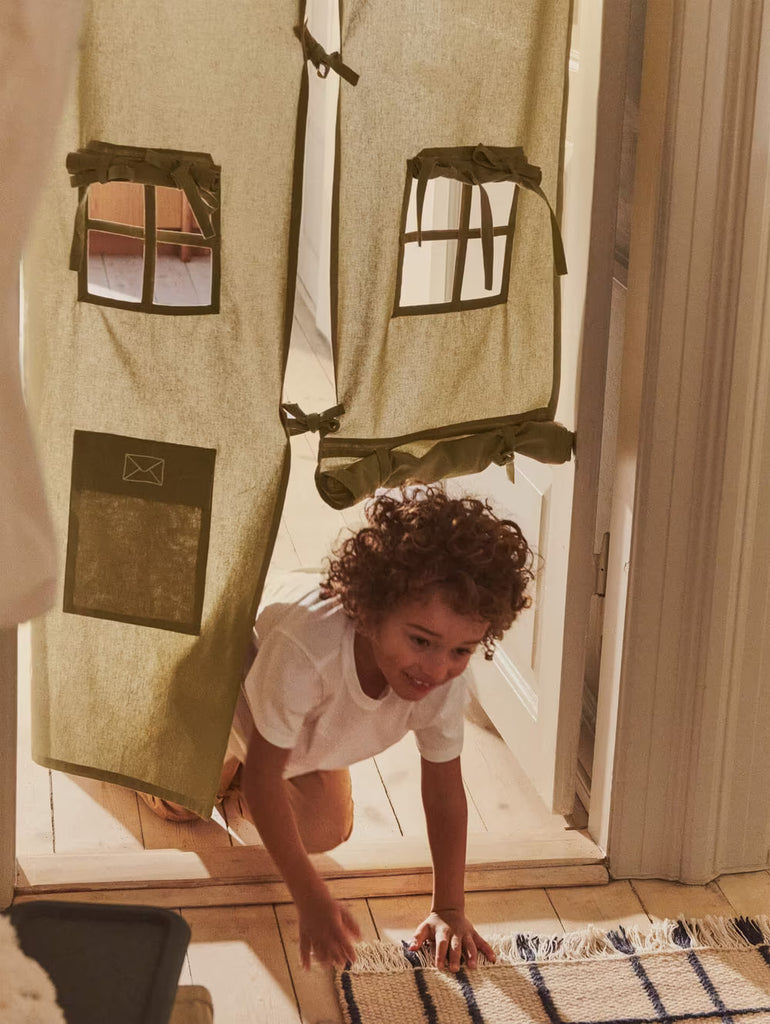 A young child with curly hair crawls on the floor, smiling, through a doorway adorned with the Ferm Living Settle Door Curtain in Elm Green—crafted from recycled cotton and designed to look like a charming house, complete with delightful window and mailbox accents.