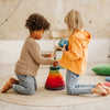 Two young children kneel on a round rug, playing with a Grimm's Giant Geometrical Stacking Tower. One child, in a beige sweater, places a ring on the tower while the other, in an orange hoodie and glasses, holds another ring. The background features a soft, neutral-toned room with calming decor.