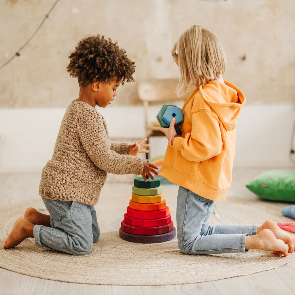 Two young children kneel on a round rug, playing with a Grimm's Giant Geometrical Stacking Tower. One child, in a beige sweater, places a ring on the tower while the other, in an orange hoodie and glasses, holds another ring. The background features a soft, neutral-toned room with calming decor.