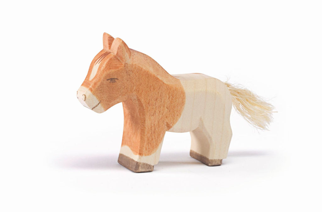 An Ostheimer Shetland Pony Colt—small, handcrafted, and featuring light brown and cream tones with a short yarn tail—stands against a white background, showcasing the charm of wooden toys.