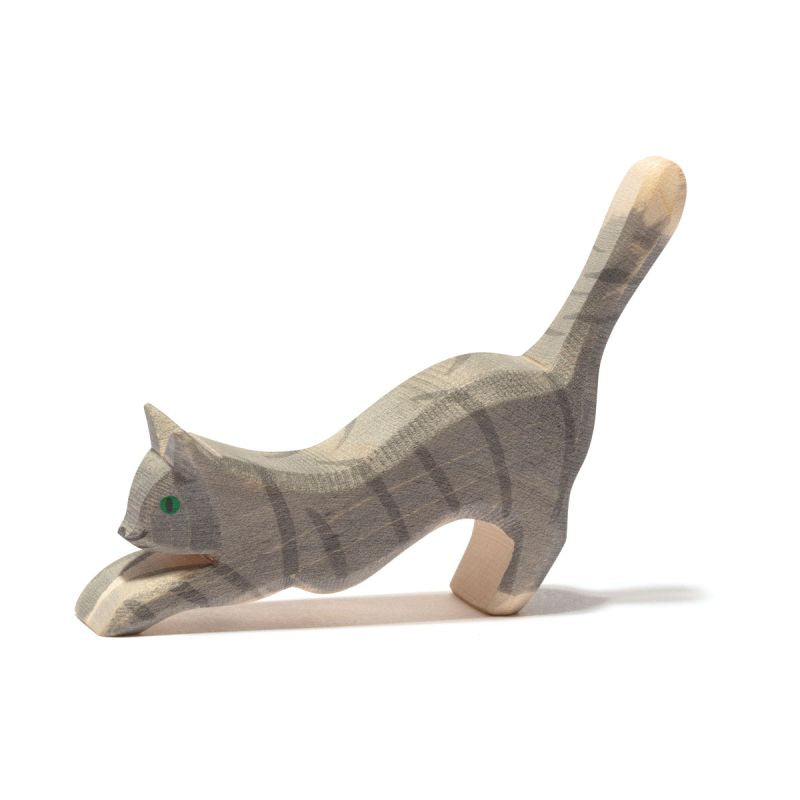 The Ostheimer Gray Cat, Jumping is a small, carved wooden figure that evokes the charm of handcrafted wooden toys. It depicts a cat in a playful, stretching position with its tail raised high and front paws extended forward. The cat's subtle green eyes and faint grey stripes on its back and tail enhance its lifelike appearance.