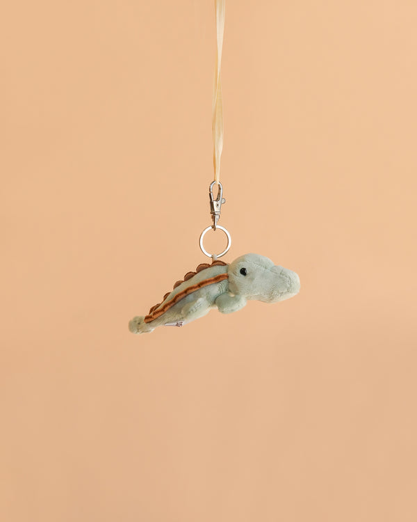 The Bukowski Maximus Alligator Bag Charm, a small plush alligator keychain featuring a metal clip and ribbon, hangs against a plain beige background—making it the perfect backpack accessory.