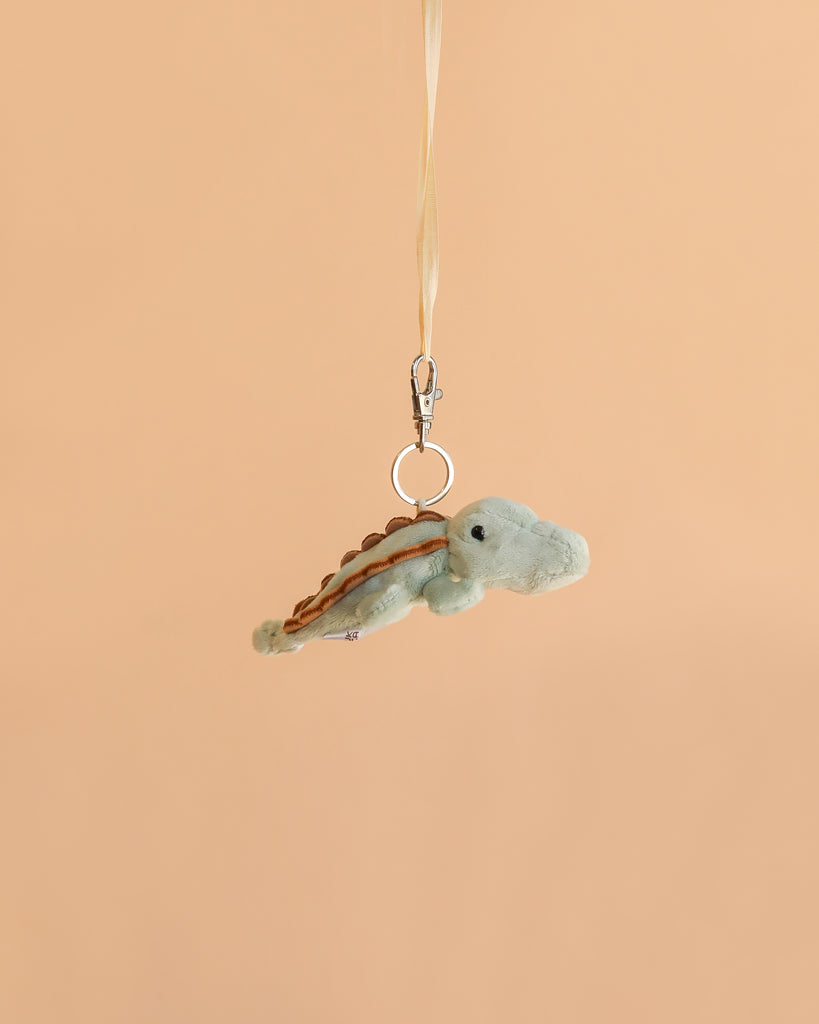 The Bukowski Maximus Alligator Bag Charm, a small plush alligator keychain featuring a metal clip and ribbon, hangs against a plain beige background—making it the perfect backpack accessory.