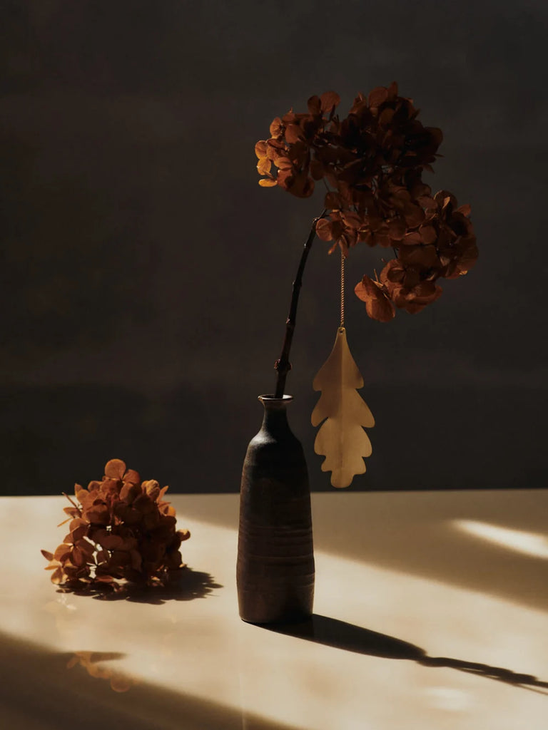 A Ferm Living Oak Leaf Ornaments - Set of 4 - Brass hangs from a dried hydrangea stem in a narrow vase.