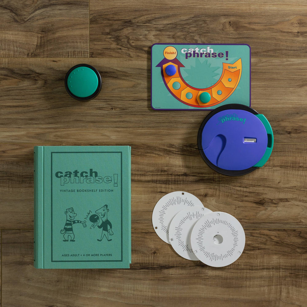 Board Game: Catch Phrase Vintage Bookshelf Edition shown on a wooden surface with a green book-shaped box, circular game unit, buzzer, two white discs, and a green-orange scorecard.
