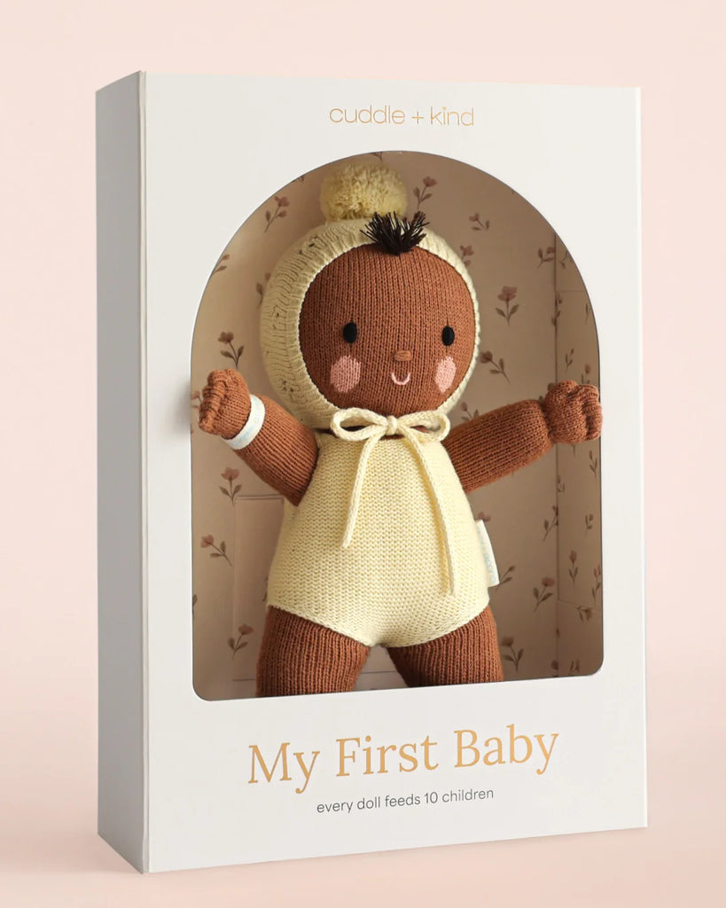 The Cuddle + Kind My First Baby (Buttercup) is a hand-knit baby doll in a brown and cream outfit, made from natural cotton yarn. Each fair trade doll feeds 10 children and ships in one week. Subtle floral patterns accent its box.