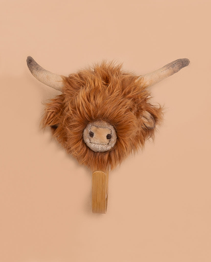 The Coat Hanger - Highland Cow is a plush wall-mounted decoration with long horns, shaggy brown fur, and a smiling face, doubling as an adorable coat hanger against a plain beige background.