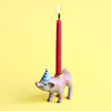 A whimsical hand-painted Pig Cake Topper shaped like a pink rhinoceros with a blue and white striped party hat, holding a lit red candle, against a yellow background.