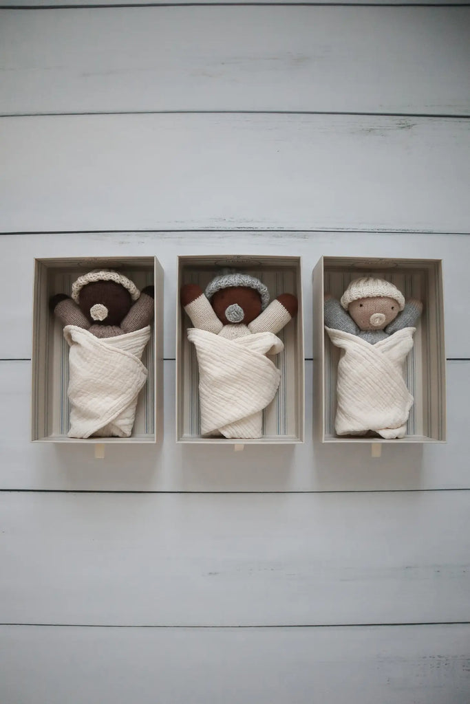 Three Hand-Knit Baby Doll - Caramel wrapped in organic cotton swaddles lying in separate wooden boxes against a light-colored wooden background.