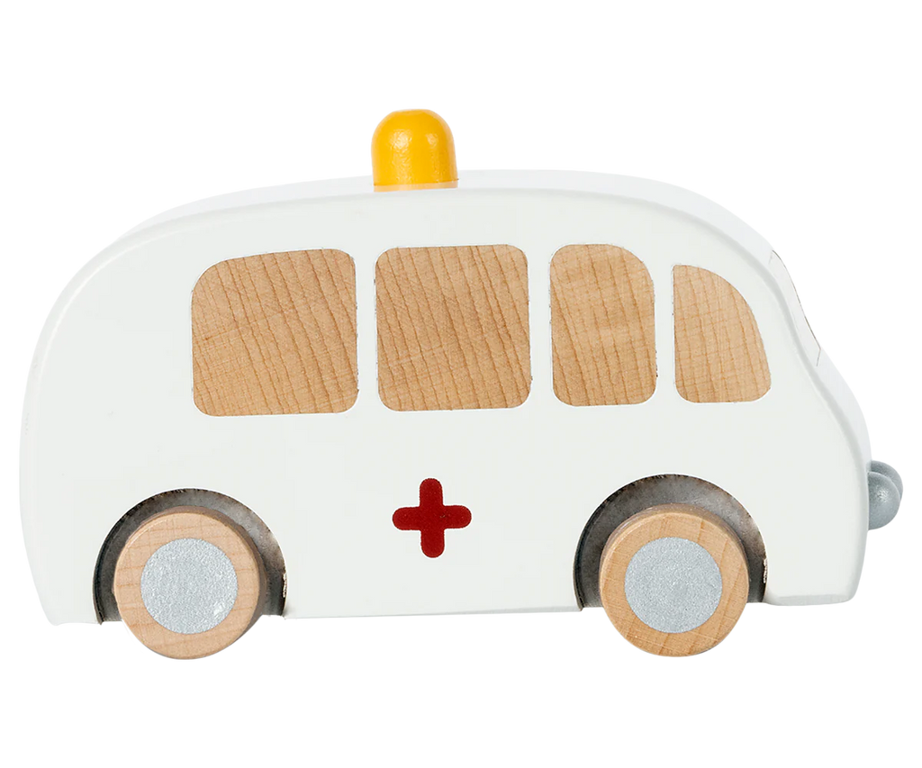 The Maileg Wooden Ambulance boasts a white body with natural wood window accents, a red cross on the side, gray wheels, and is made from solid wood. It features a yellow light on top and a pull-back function for endless play.