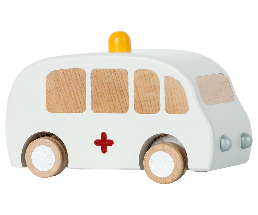 The Maileg Wooden Ambulance is a charming toy featuring solid wood construction, white paint with a red cross on the side, four wooden wheels, and a small yellow light on top. Its natural wood color windows and push or pull-back function offer delight.