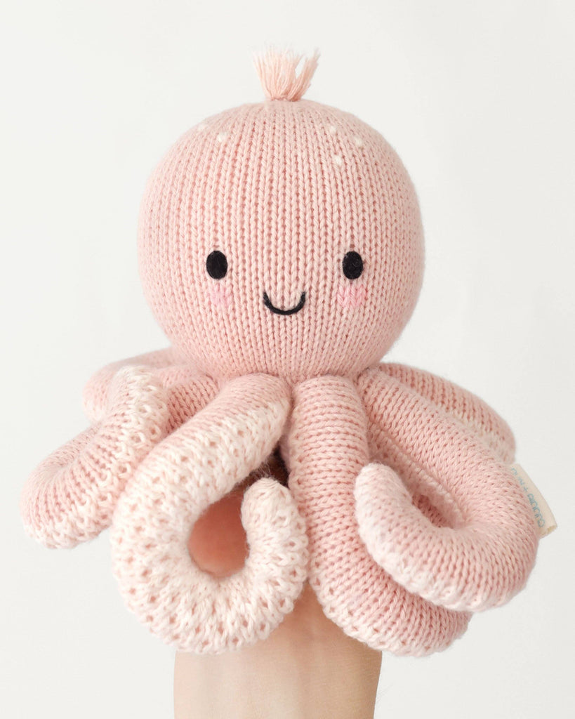 A hand holds the Cuddle + Kind Baby Octopus (Soft Coral Pink), an heirloom-quality, hand-knit plush toy with a smiling face, black eyes, and light pink tentacles, crafted from soft Peruvian cotton yarn against a white background.