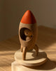 The Sabo Concept Rocket with Bear features a wooden rocket with an orange tip, a small bear inside, and stands on a round base—perfect for imaginative play in warm sunlight.