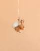 The Bukowski Hera Bunny Bag Charm is a small, soft plush bunny keychain with tan fur and a white fluffy tail, hanging from a cream-colored ribbon against a plain peach background—perfect as an adorable backpack accessory.