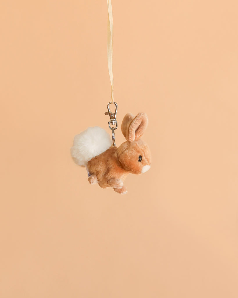 The Bukowski Hera Bunny Bag Charm is a small, soft plush bunny keychain with tan fur and a white fluffy tail, hanging from a cream-colored ribbon against a plain peach background—perfect as an adorable backpack accessory.
