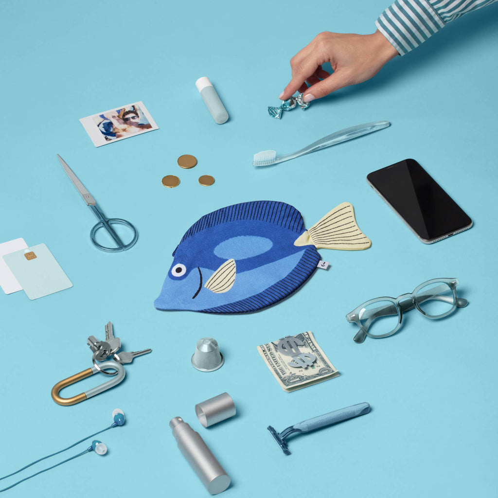 A hand reaches for a wrapped candy among various items, including the Surgeon Fish Pencil Case, a smartphone, glasses, money, scissors, toothbrush, coins, and toiletries—all laid out on a light blue background.