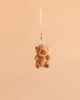 A small, brown Bukowski Theo Capybara Bag Charm hangs from a beige ribbon against a plain, light peach background.