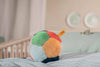 A Steiff Multicolored Plush Ball with Rattle and yellow tag rests on a light blue-striped bed in a softly lit bedroom. Easy to grip, this plush ball is near a wooden crib and two decorative plates in the background.