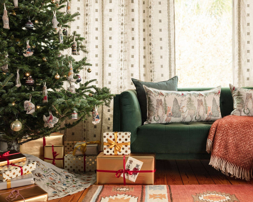 A cozy living room showcases a decorated Christmas tree adorned with shimmering ornaments, including a Yeti with Candy Cane ornament, standing beside a green sofa. The floor is embellished with gifts wrapped in gold and white paper. The sofa is accented with festive pillows and a warm pink throw blanket, as light softly filters through patterned curtains.