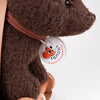 Close-up of a brown Pull-Along Raoul plush bear with a round tag on its neck. The tag features a cartoon fox face, the text “EGMONT,” and the phrase “a world of imagination.” The plush toy bear is set on wooden wheels.