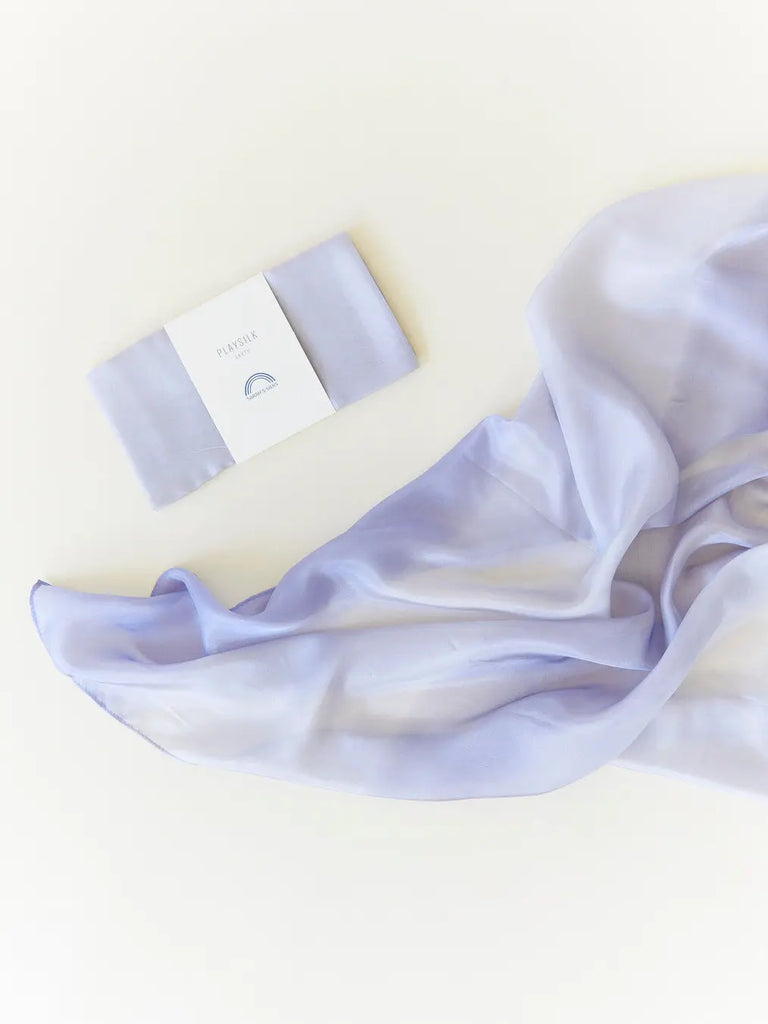 A delicate lavender Sarah's Silk Earth Playsilk - Stone gracefully strewn next to a small white product box labeled "pure silk" on a light background. The composition conveys elegance and softness, inviting imaginative play.