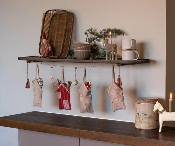 A wooden shelf displays festive decor, while below hangs the Maileg Christmas Street Advent Calendar, featuring five numbered fabric pouches with embroidered details and filled with small Christmas decorations.