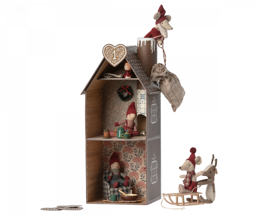 A decorative Christmas-themed Maileg Cardboard Two-Story Gingerbread House with three levels featuring festive dolls and ornaments such as a wreath, gingerman, and a gnome. A sled rests outside the house, set against