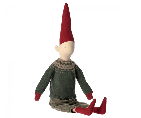 A Maileg Christmas Pixy (Size 5) with a pointed red hat, green sweater, brown pants, and red shoes, seated on an invisible surface against a black background, designed as an exclusive Christmas decoration.