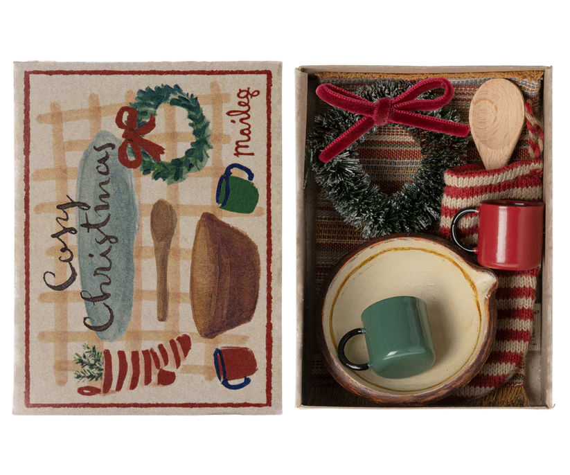 The Maileg Cosy Christmas Set is a delightful gift box featuring charming Christmas accessories, including a wooden spoon, woven fabric, and a decorative wreath adorned with a red ribbon. Inside the box, you'll find a green mug and a red mug. The lid is beautifully illustrated with mugs and wreaths, accompanied by the cheerful text "Cozy Christmas" in a festive design.