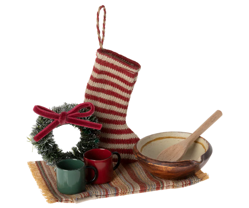 The Maileg Cosy Christmas Set includes a delightful arrangement featuring a red and white striped Christmas stocking, a small wreath with a red bow, two mugs (one green, one red), and a wooden spoon in a ceramic bowl, all beautifully placed on a striped cloth mat with charming Christmas accessories.