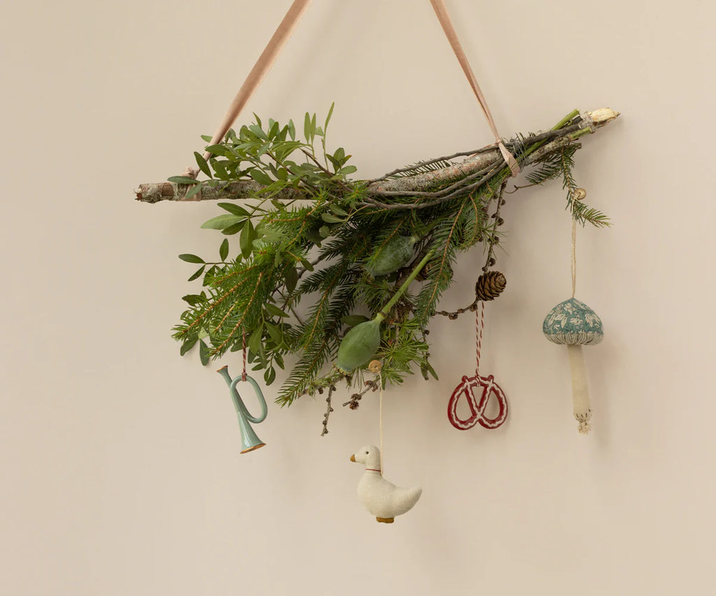 A decorative branch hanging on a wall showcases green foliage, a pine cone, a large letter "P," and delightful Maileg ornaments including a mint-colored trumpet-shaped metal ornament.