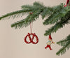 A close-up of the Maileg Christmas Metal Ornament - Pretzel - Dot, a hand-painted red pretzel with white dots, displayed on a pine branch. The unique ornament adds charm to your Christmas tree against a plain beige background.