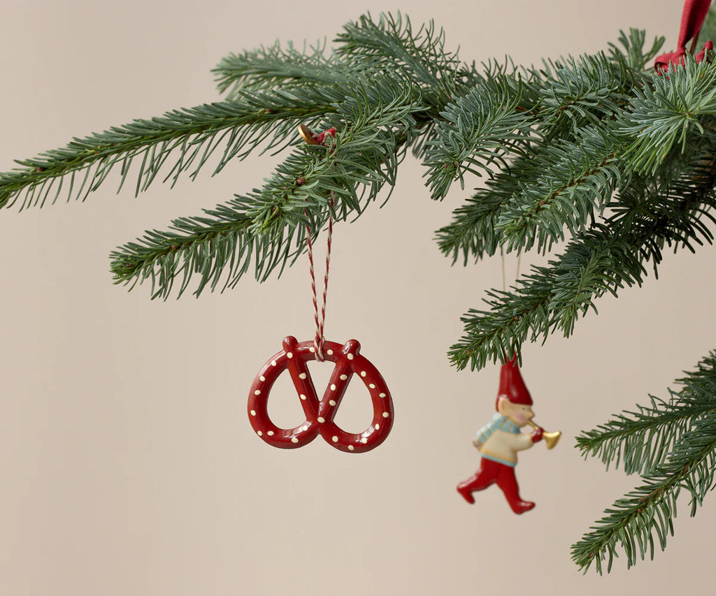 A close-up of the Maileg Christmas Metal Ornament - Pretzel - Dot, a hand-painted red pretzel with white dots, displayed on a pine branch. The unique ornament adds charm to your Christmas tree against a plain beige background.